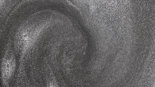 Mesmerizing close-up of silver paint swirling continuously to form a deep vortex while glittering particles flow and rotate to reveal a metallic sandy texture on abstract background