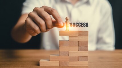 A persons hand carefully places a small wooden figure on the highest block of a rising wooden staircase beside the word SUCCESS with warm light glowing on a wooden table