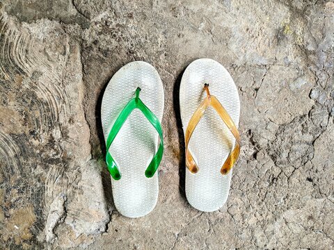 Top view of worn-out flip-flops with green and orange straps on a cracked concrete floor.