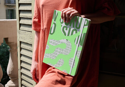 Holding a Mockup Notebook by the BalconyWoman stands on a balcony holding a blank vertical notebook. PSD template mockup.