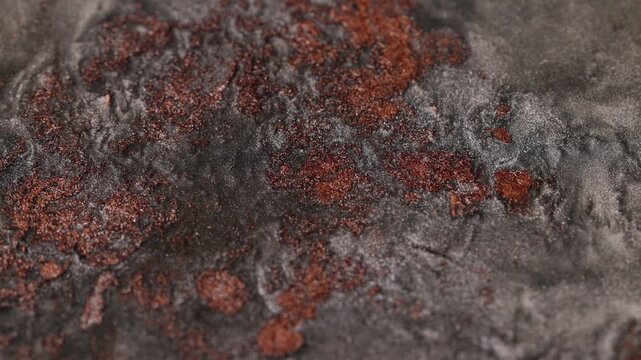 Macro close-up of dark titanium metallic fluid bubbling and shifting slowly to reveal patches of reddish copper glitter particles creating a rough cratered texture resembling cooling lava 
