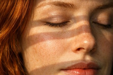 Naklejka premium Close-up of freckled woman face with closed eyes and soft sunlight creating gentle shadow pattern.