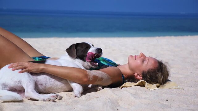 Beautiful young woman sunbathing and relaxing on a tropical white sand beach while her cute dog sleeps peacefully on her chest, enjoying a sunny summer day on a seaside vacation together