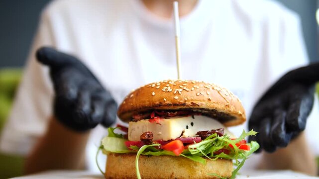 Professional chef in black gloves serving a tasty vegan burger with tofu cheese, fresh vegetables, and sesame seed bun in a restaurant, a perfect example of modern, healthy fast food