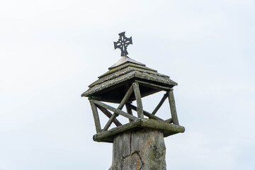 weathered wooden folk shrine on timber pillar featuring decorative iron cross against bright overcast sky