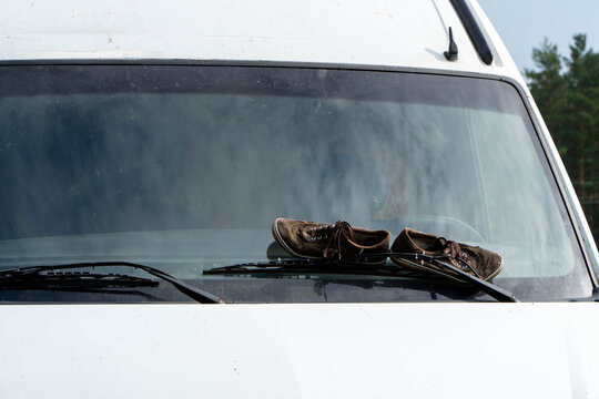 Wide view of brown canvas sneakers drying on windshield of white camper van held by wiper blades in outdoor rural setting.