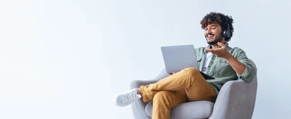 Fototapete Altes Krankenhaus Beelitz A young man sits comfortably in a chair, wearing headphones and smiling. He uses a laptop for an online meeting. The background is simple and bright, creating a relaxed setting.  © Prostock-studio