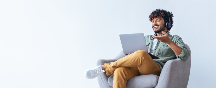 A young man sits comfortably in a chair, wearing headphones and smiling. He uses a laptop for an online meeting. The background is simple and bright, creating a relaxed setting.