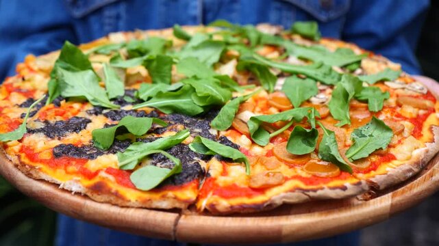 Unrecognizable person presenting a delicious homemade vegan pizza on a wooden cutting board, topped with fresh arugula, sausage slices, mushrooms, and black olive pate for a healthy meal