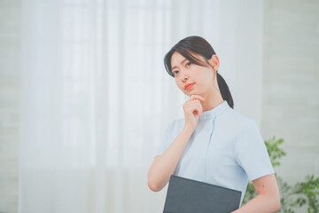 明るく清潔感のあるクリニックで考えるポーズをする20代女性看護師