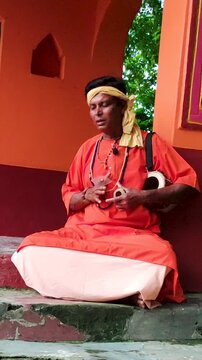 A serene folk singer, dressed in an orange robe, singing with a string instrument, sitting alone on the temple veranda&mdash;a peaceful portrait of devotion, tradition, and spiritual calm in rural India.