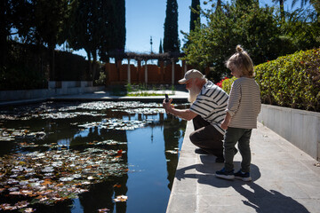 A tender moment shared between a grandfather and his grandson by the peaceful pond, with the grandfather capturing the beauty of the water lilies on his phone while the child enjoys the view