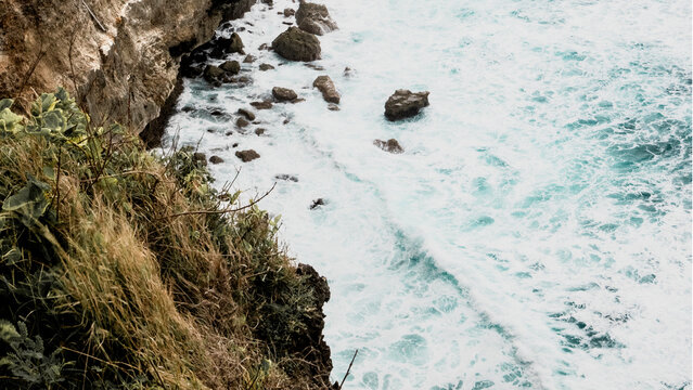Close-up view of powerful ocean waves breaking against rugged limestone cliffs along the Uluwatu coastline in Bali, Indonesia. Textured rock formations and foaming sea water.