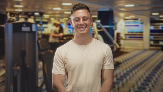 Man with glasses wearing beige tshirt, smiling and standing by exercise machines in a studio gym interior with patterned carpet; confidence fitness wellness.