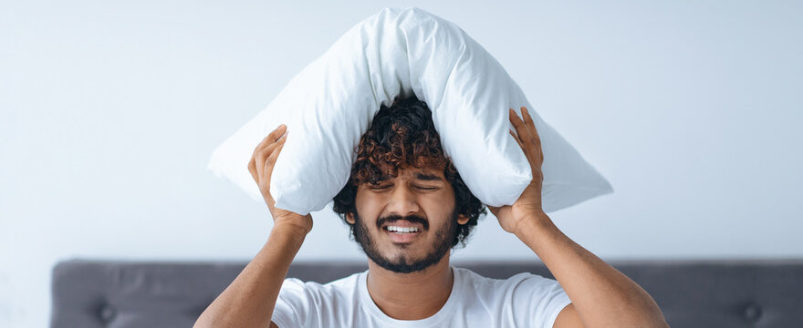 A man is sitting on a bed with a pillow pressed against his head. He appears to be frustrated or overwhelmed. The setting is a simple bedroom with light colors.