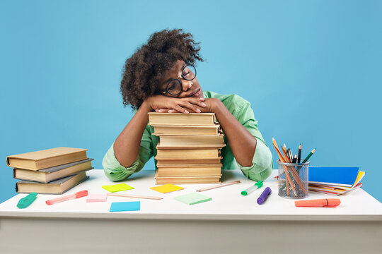 Exhausted black male student sleeping on desk on stacks of books, being tired while preparing for exam on blue studio background. African american teenager feeling overworked while studying