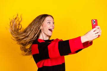 Cheerful young woman taking a selfie with a red smartphone against a vibrant yellow background,...