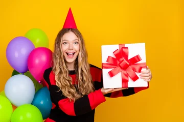 Fotobehang Onderzeeër Excited young woman in festive outfit holding a gift box against colorful balloons and vibrant yellow background  © deagreez