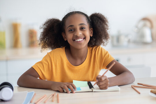 Cheerful inteligent african american teen girl in casual doing homework, sitting at cozy kitchen at home, taking notes or making plan, using pencils and notepad, smiling at camera, closeup portrait