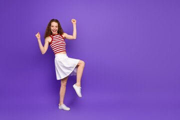 Young woman jumping with joy in red striped sleeveless top and white skirt against purple background for fashion lifestyle and advertising imagery