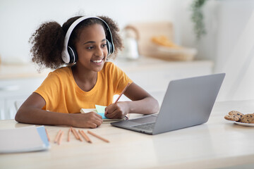 Cheerful black girl studying online, using laptop and headset at home, copy space. Cute african...