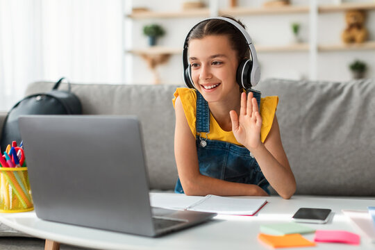 Remote Learning Concept. Portrait of positive girl in headset sitting at desk on couch in living room, using computer having virtual chat, waving hand to webcam greeting teacher and classmates