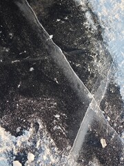 Close up texture of cracked black ice covered with white snow powder. Abstract winter background of frozen water surface with deep fissures