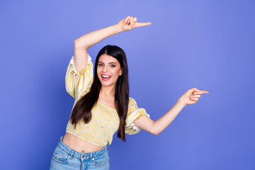 Fun stylish young woman posing in yellow blouse and jeans smiling pointing purple background for...