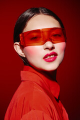 Fashion portrait of a woman wearing transparent red sunglasses and glossy red lipstick. Her makeup...