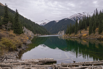 Scenic view of a calm mountain lake surrounded by evergreen forest and alpine slopes, with snow-capped peaks in the background and reflections on the water.