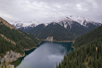 Aerial view of a calm mountain lake surrounded by evergreen forest and alpine slopes, with snow-capped peaks in the background and reflections on the water.