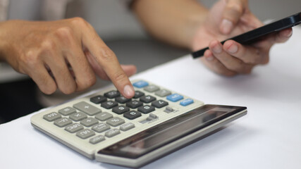 Close up of professional hands simultaneously using a grey office calculator and holding a black smartphone. Concept of multitasking, financial calculation, tax planning, and mobile accounting