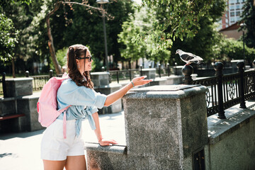 Young woman with pink backpack smiling as she feeds a pigeon in a sunny outdoor park in the city...