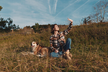 Obraz premium Woman and dog in an open field under a blue sky, enjoying nature and sunshine. Casual plaid shirt, jeans and boots as she sits and waves toward the bright horizon.