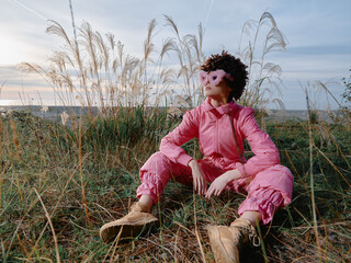 Obraz premium Outdoor fashion scene with a model wearing a pink jumpsuit and mask, seated in tall grasses during golden hour, presenting bold style and nature inspired calm.