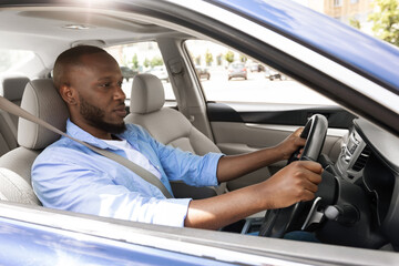 Confident Professional Driver. Side view profile portrait of young African American man sitting in a car on driver's seat. Focused black guy riding in the city, holding hands on steering wheel © Prostock-studio