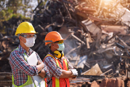 Two manual workers wearing face masks and protective workwear standing side by side in a scrap metal yard, Thailand