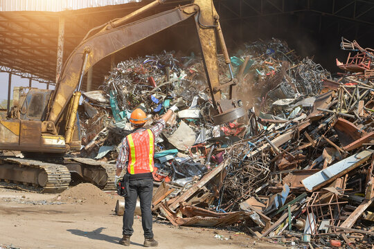 Rear view of a worker at a metal scrap yard pointing at an excavator with a magnet, Thailand