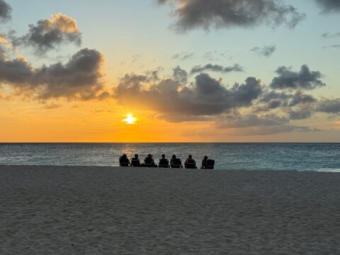 Rear view silhouette of a row of seven people sitting on Eagle beach watching the sunset, Aruba