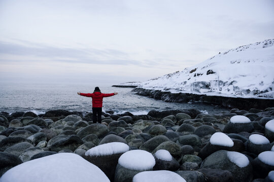 Rear view of a man in a puffer coat standing with his arms outstretched on Barents Sea coast in winter, Teriberka, Kolsky District, Murmansk Oblast, Russia