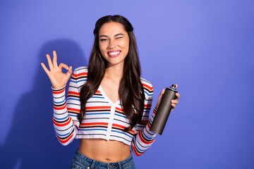 Young female model smiling and winking while holding a spray bottle in front of a purple background...