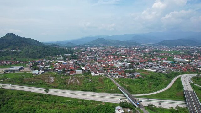 Aerial drone footage of the whole big city of Sumedang, in Java island, Indonesia, inside it's valley of mountains, with a motorway interchange in the front, in a cloudy atmosphere