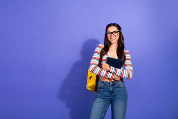 Young smiling brunette student with glasses stands against violet background holding notebook and...