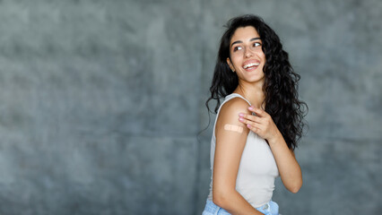Fototapeta premium Coronavirus vaccination, viral disease prevention concept. Happy young woman showing band aid on her arm, looking aside at free space on grey background, banner design