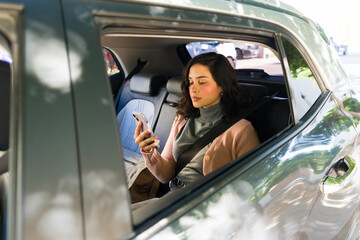Latin woman enjoying a rideshare service, checking messages on her mobile phone while traveling in...