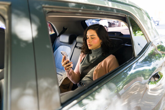 Latin woman enjoying a rideshare service, checking messages on her mobile phone while traveling in the back seat of a car