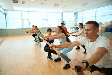 People doing squats with female trainer holding gymnastic sticks in fitness club