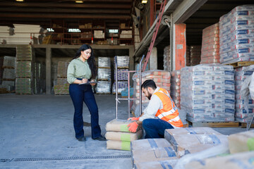 Woman manager checking inventory and worker moving cement bags in a construction supply warehouse for business to business logistics