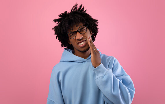 Unhappy black teenager suffering from tooth ache, having gum problems, touching his cheek on pink studio background. African American adolescent suffering from dental pain, having sensitive teeth
