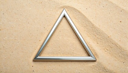 A silver triangle frame sits on a sandy surface with light rippling patterns, creating depth and texture. The geometric shape contrasts the background